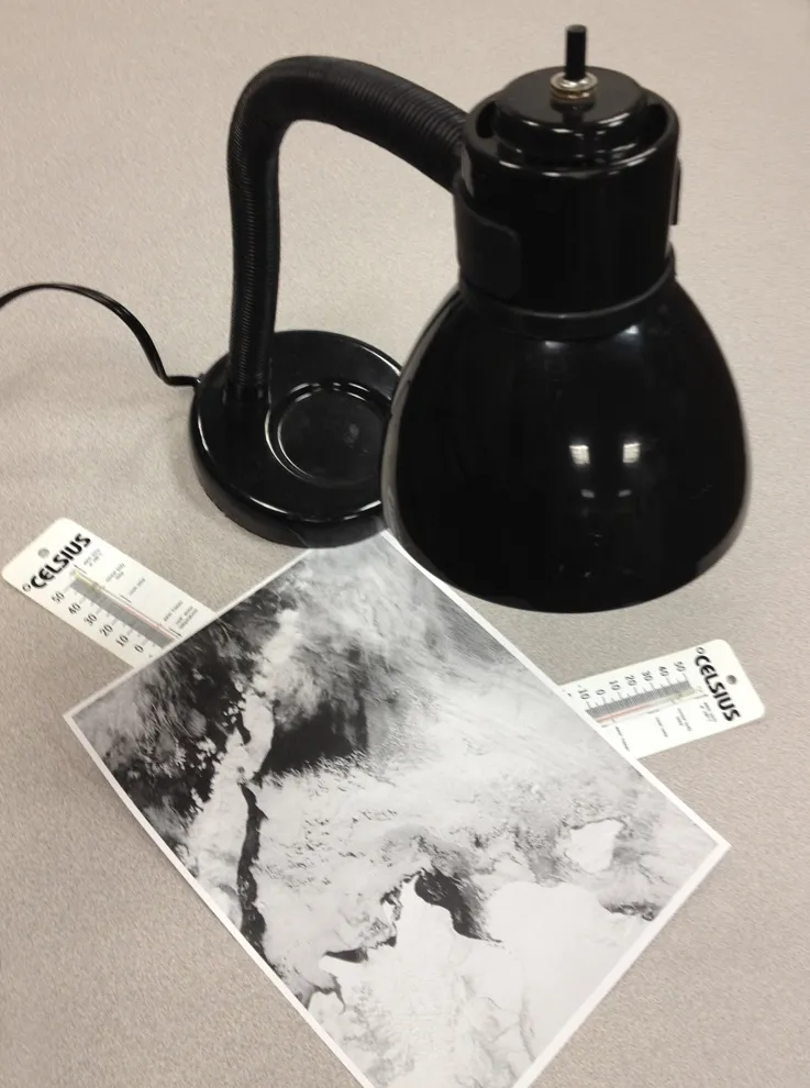 Setup for the albedo activity. A lamp shines on a black and white image. Two thermometers measure the temperature beneath the black areas of the image and the white areas of the image.