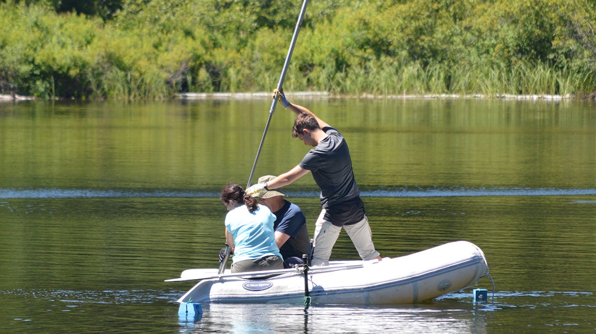 Three scientists in a small inflatable boat on a lake take a sediment core from the lake bottom.