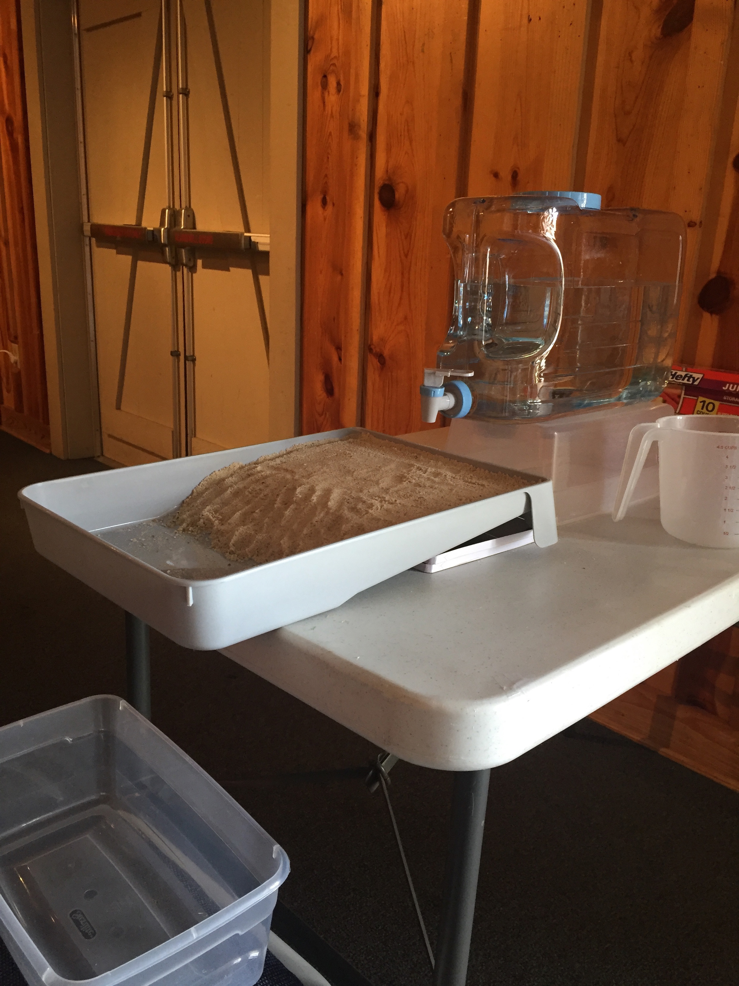 A photo of the stream table set-up, showing the water jug positioned over one end of the stream table tray, and a bin to catch drainage water at the bottom.