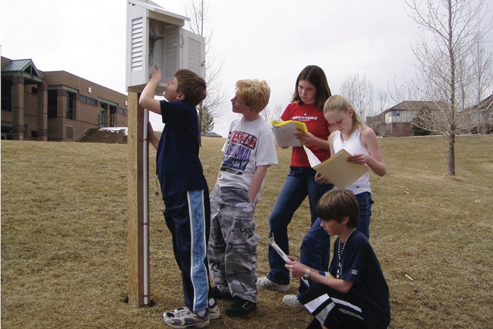 A group of students are taking measurements from the weather instruments inside the instrument shelter, which is installed on a post near their school. 