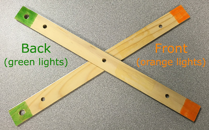 Simple UAV model made from wooden rulers. Two rulers are connected at the middle in an X form. Two ends of the X are painted green to represent the green lights on the back of a UAV. The other two ends of the X are painted orange to represent the orange lights on the front of a UAV