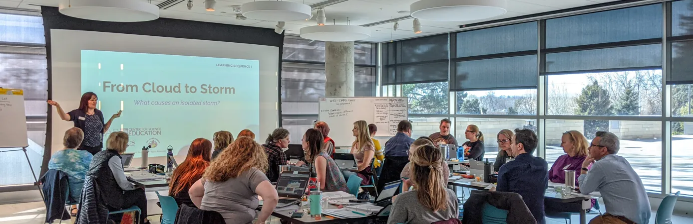 A group of seated educators look towards a screen and a presenter during a learning workshop.