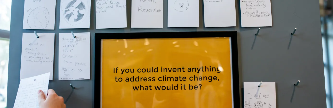 A sign on a wall reads "If you could invent anything to address climate change what would it be?" There are cards hanging around the sign that have ideas for climate solutions written on them.