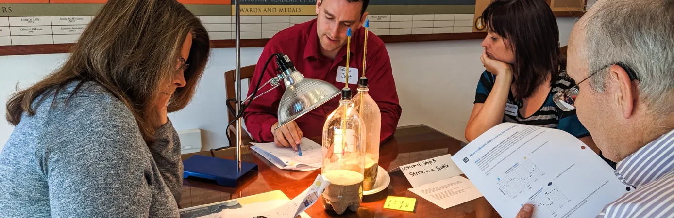 Four teachers sitting around a table are recording data about an experiment with a lamp shining on two bottles filled partially with sand and water. Thermometers are inserted in each bottle.