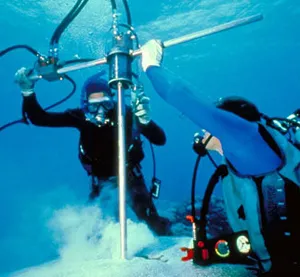 Two scientists underwater in scuba gear hold a drill to coral on the seafloor.