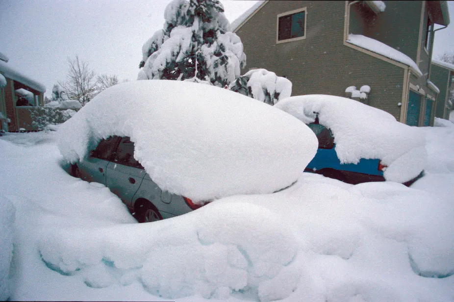 A thick blanket of snow covers two cars parked in front of a house