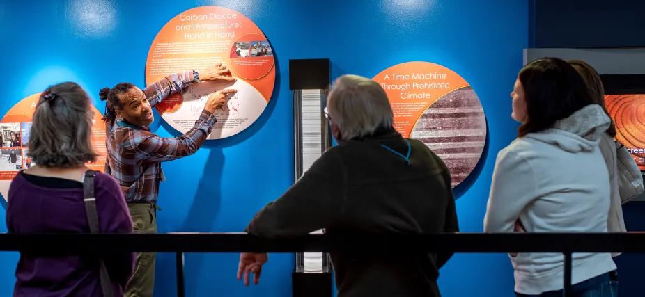 A photo of a tour guide pointing at an exhibit panel in the NCAR Climate Exhibit. Four people standing around the tour guide attentively. 