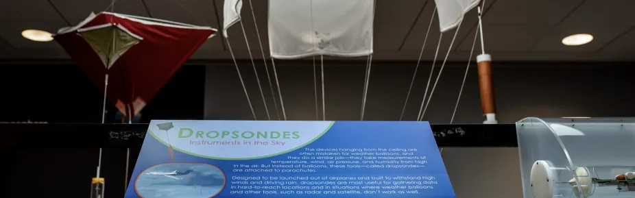 An exhibit sign titled “Dropsondes” in front of four dropsondes that are hanging from the ceiling.