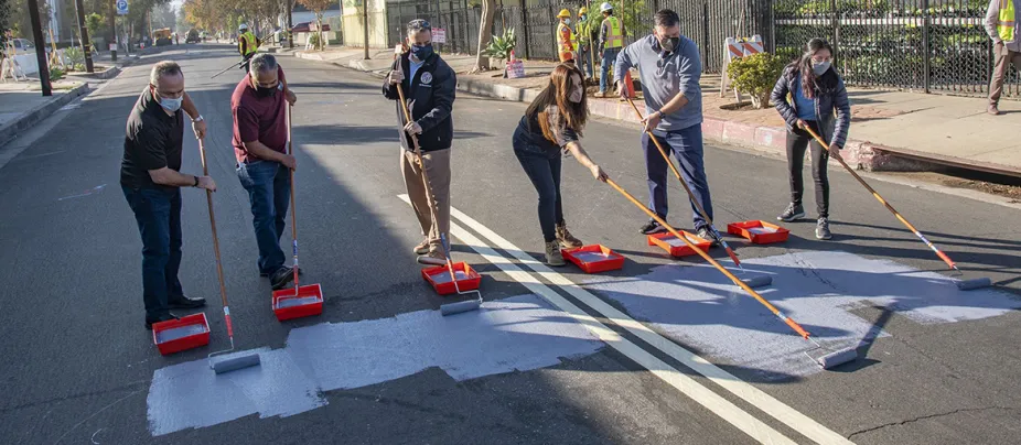 Six people stand in a Los Angeles street holding paint rollers. They are painting the pavement with a grey paint, which will reflect energy from the Sun.