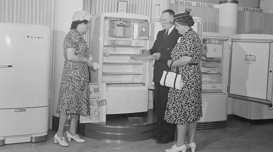 In this black and white 1941 photo, two white women wearing patterned summer dresses, white shoes, and hats look inside a refrigerator in a department store showroom as a salesman in a dark suit and tie shows them an ice tray from the freezer.