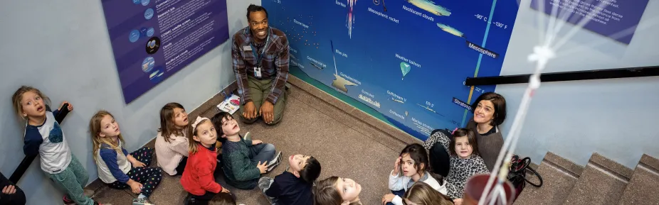 A group of students sitting around an educator and all looking up in wonder at something above them.