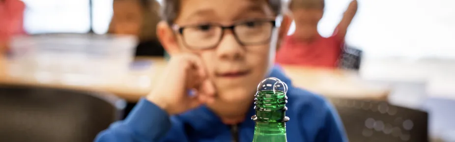 A small soap bubble protruding from a green water bottle. In the background is a student watching curiously.