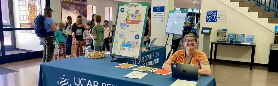A staff member sitting behind a table smiling at the camera. Behind her is an educator surrounded by a group of students and parents.