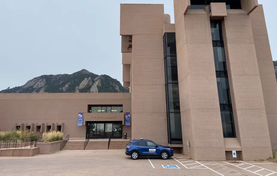 A photo of the front entrance to the Mesa Lab. The entrance has text above the doors that read “National Center for Atmospheric Research, Sponsored by the National Science Foundation.” There are two blue banners on either side of the entrance that have the text “Discover” and “Explore” on them. This photo shows Tower 1 of the building with three accessible parking spots in front and a blue car parked in one of them. Peeking out behind the building is Bear Peak with a bit of the mountains below it.