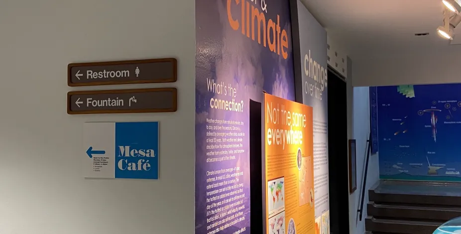 A photo showing three wall signs at the Mesa Lab; one with an arrow that says “Restroom”, one with an arrow that says “Fountain”, and the final one with an arrow that says “Mesa Café”. In the background of this photo is the end of the weather exhibit with the NCAR Theater and the stairs up to the Climate exhibit. 