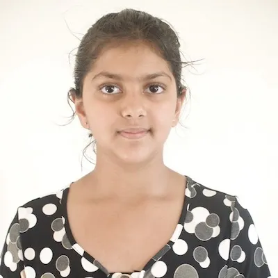 A photo of Madhvi smiling at the camera. Madhvi has brown skin, dark hair tied up behind her head, and is wearing a polka-dot shirt.