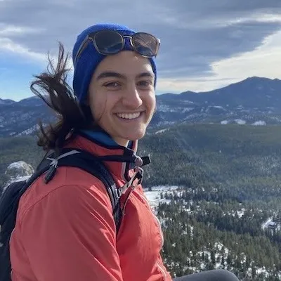 A photo of Maya smiling at the camera. Maya has brown skin, dark hair which is blowing in the wind, and is wearing a blue beanie, sunglasses, and a red windbreaker.