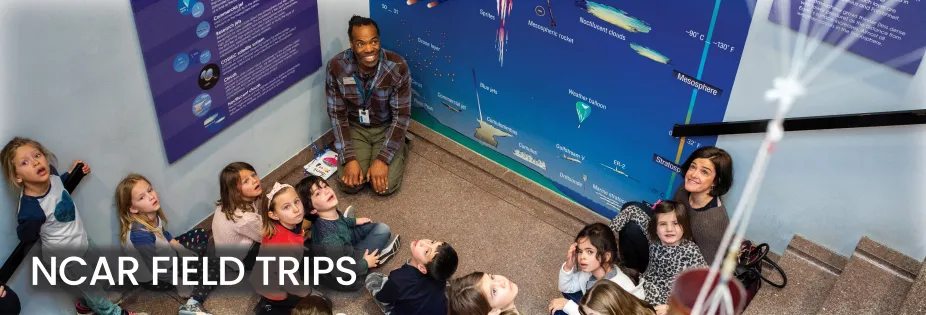 A photo shooting down the stairs of an educator sitting on the stair landing with a group of students looking up towards the top of the stairs. A mural of the layers of the atmosphere is behind them.