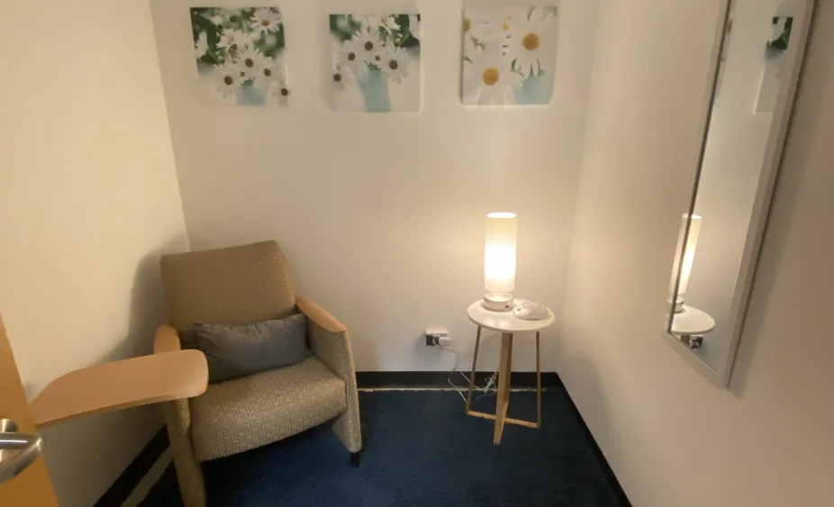 Photo of a calmly lit room with a chair, pillow, and table in one corner and a table, lamp, and noise machine in the other corner. There is an outlet on the wall opposite the door.