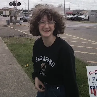A photo of Phoebe smiling at the camera. She has light brown curly hair, pale skin, and is wearing rounded glasses and a navy blue long sleeve t-shirt.