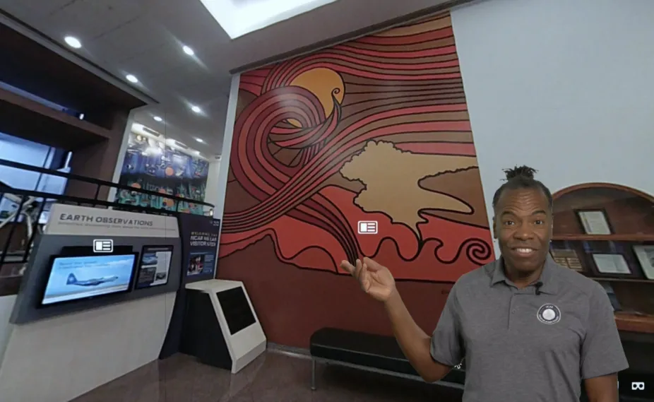 a tour guide stands in front of a digital image of the Mesa Laboratory Visitor Center Lobby pointing at a mural