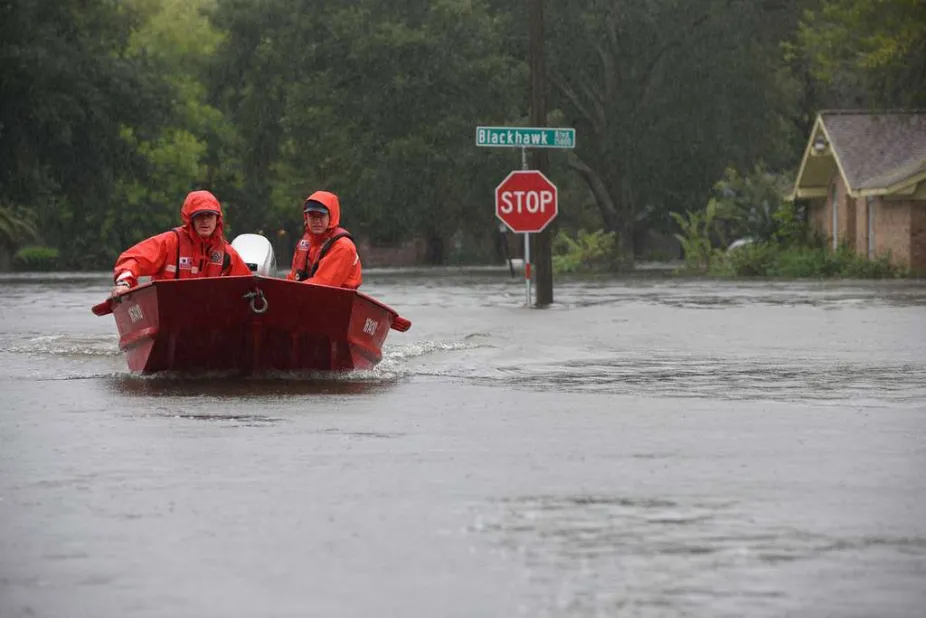 Two people in red coats in a small boat in a flooded street. A street sign and stop sign are above the water level.   