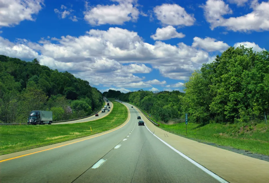 Small cumulus clouds dot the sky above a road through green forested land