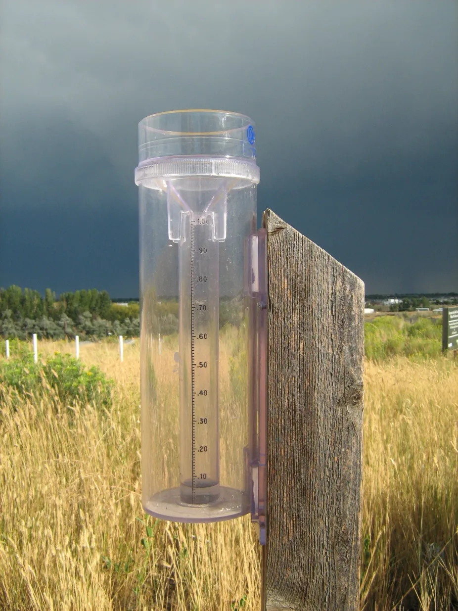 plastic rain gauge on a wood post in a grassy field under dark storm clouds