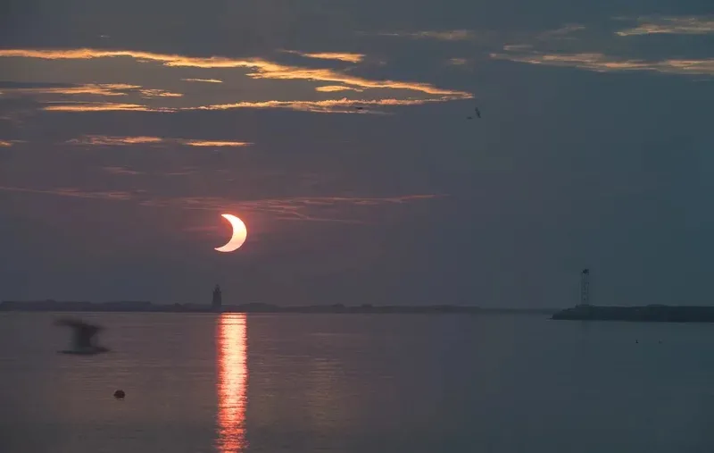 A partial solar eclipse in a darkened sky over the water. The eclipse is reflected in water and the Sun appears as crecent-shaped, partially obscured by the Moon.
