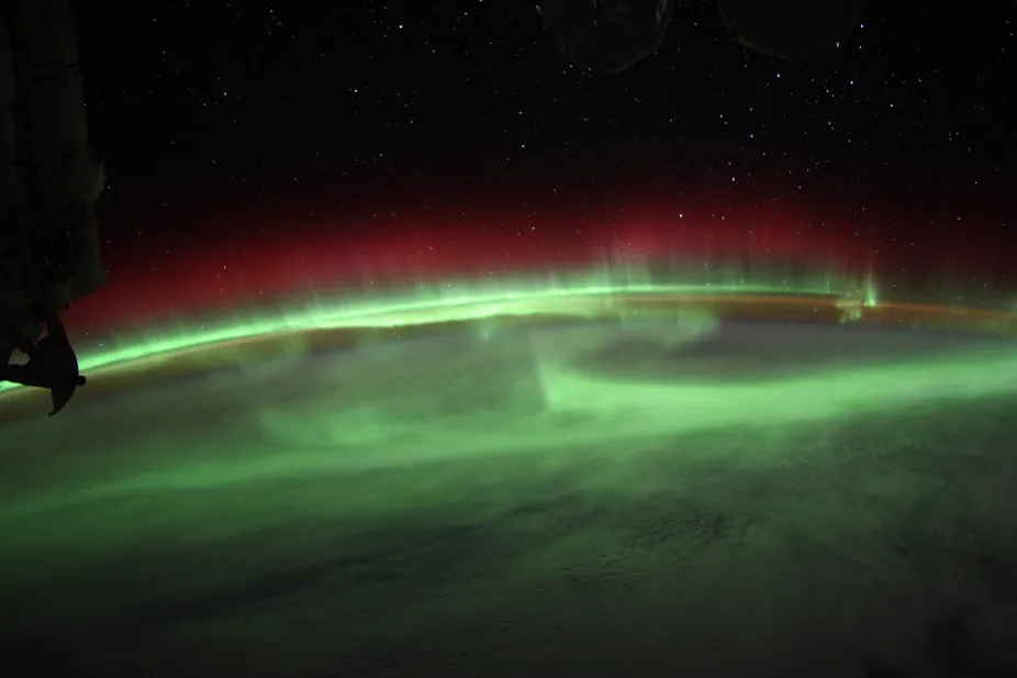 A glowing band of lights extends across the Earth's atmosphere, with a bit of the International Space Station visible from the left side of the image.