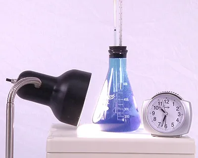 A lamp facing a flask with rubber stopper that holds a tube and thermometer, and a clock