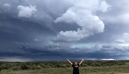 A photo of someone standing with their arms raised excitedly and a dark storm is looming behind them.