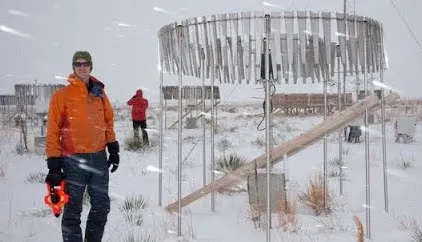 A photo of a scientist in an orange snow coat standing next to a scientific device as snow falls all around. 