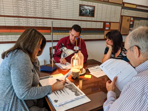 Four teachers sitting around a table are recording data about an experiment with a lamp shining on two bottles filled partially with sand and water. Thermometers are inserted in each bottle.