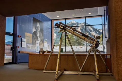 A photo of the the White Light Coronal Camera, designed and built at NCAR in 1966. The camera is around 6 feet tall and 8 feet wide. Behind the camera is the architecture exhibit with a large photo of I.M. Pei overlooking a view of the mountains in the Boulder foothills.