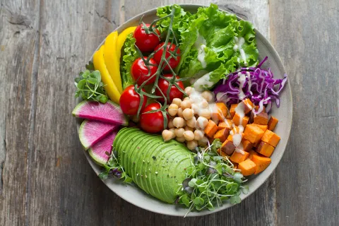 A plate of salad with sliced avocado, lettuce, tomatoes on the vine, sliced bell pepper, chickpeas, cubes of sweet potato, shredded cabbage, and sprouts, with dressing drizzled over the top.